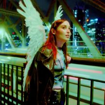 Hatchie, wearing angels wings, standing by an empty road at night and looking away from camera with high-rise buildings in the background