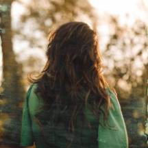 The torso of a woman with long brown hair looking away from camera. The background is sky and blurred trees