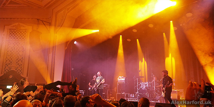 Widescreen shot of gig with the stage to the right and Ross crowdsurfing to the left. Stagelighting is orange/yellow.